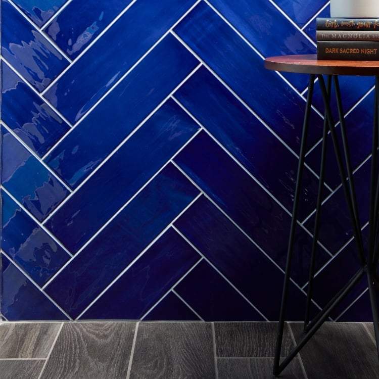 4x16 Gioia Navy colored tile set in a herringbone pattern on a wall close up abutting a wood-look floor.